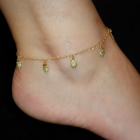 A person's ankle features a captivating anklet from the Owl Charms collection, filled with wisdom and intuition. It is delicately adorned with small dangling hearts as lucky charms against a black background.